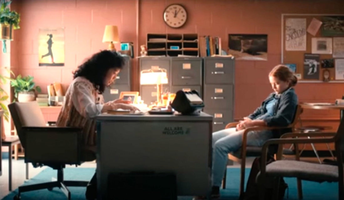 Ms. Kelley at her school desk, Max seated across from her, in front of the filing cabinets